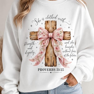 Può includere: Una felpa bianca a girocollo con la grafica di una croce in legno decorata con un grande fiocco floreale rosa. Il testo recita: She is clothed with strength and dignity, and she laughs without fear of the future. PROVERBS 31:25.