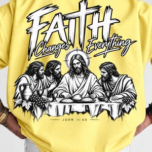 Pode incluir: Uma t-shirt amarelo-brilhante com uma impressão gráfica a preto e branco nas costas. O design retrata uma cena de quatro figuras numa mesa com o texto "FAITH Changes Everything" acima e "JOHN 11:40" impresso abaixo da ilustração.