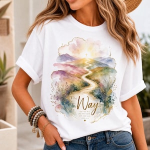 Può includere: Una t-shirt bianca con una grafica in stile acquerello raffigurante un sentiero tortuoso che conduce verso una croce su una collina. Il testo recita The Way, I am the way, the truth, and the life. John 14:6. Il design include montagne e fogliame pastello.