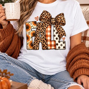 Könnte beinhalten: Ein weißes Kurzarm-T-Shirt mit dem Wort FALL in großen Blockbuchstaben. Die Buchstaben sind mit Sonnenblumen-, Leoparden-, orangefarbenen Karomustern und schwarzen Punkten verziert und mit einer großen Leoparden-Schleife versehen.