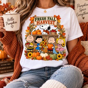 Op de afbeelding: Een wit t-shirt met een afbeelding van de Peanuts-figuren met pompoenen, zonnebloemen en herfstbladeren. De tekst luidt Fresh Fall Harvest. Het ontwerp toont Snoopy, Charlie Brown, Lucy, Linus en Sally in een oogsttafereel.