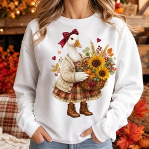 Pode incluir: Uma camisola branca de manga comprida com o gráfico de um pato branco a usar um laço vermelho, um casaco de malha creme e um vestido aos quadrados. O pato segura um cesto de vime cheio de girassóis, abóboras e folhas de outono.