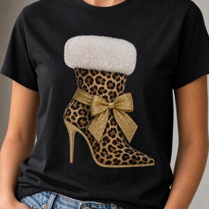 Pode incluir: Uma t-shirt preta com um gráfico de uma bota de salto alto com estampado de leopardo. A bota tem uma bainha de pelo sintético branco no topo e um grande laço dourado brilhante atado ao tornozelo. O salto e a sola são também dourados.