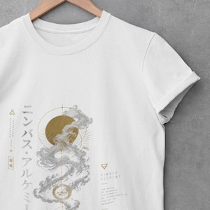 Puede incluir: Camiseta blanca con un diseño gráfico que presenta un sol dorado, nubes estilizadas y formas geométricas. También es visible el texto "Nimbus Alchemy" y caracteres japoneses. La camiseta está colgada en una percha de madera.