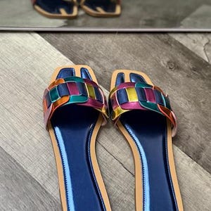 May include: A pair of colorful woven-strap sandals with a blue footbed and tan sole. The sandals feature a square toe and a metallic finish on the straps, reflecting a variety of colors. The sandals are displayed on a wooden surface.