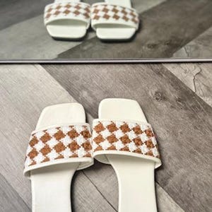 May include: A pair of white slide sandals with brown and white chequered straps. The sandals are reflected in a mirror above them, set against a wooden floor.