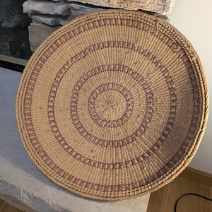 May include: A round, woven basket with a natural tan base and concentric bands of reddish-brown geometric patterns. The basket is displayed on a stone surface, showcasing its intricate design and craftsmanship. The basket's diameter is approximately 18 inches.