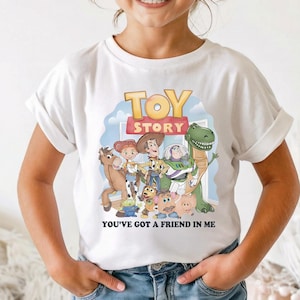 K&ouml;nnte beinhalten: Wei&szlig;es T-Shirt mit einer bunten Grafik von "Toy Story"-Charakteren. Das Shirt zeigt den Text "TOY STORY" und "YOU'VE GOT A FRIEND IN ME". Das Kind tr&auml;gt ein rosa Stirnband und eine Sonnenbrille.