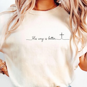 May include: A cream-colored short-sleeve t-shirt featuring the black cursive text His way is better with a small cross symbol at the end. The shirt is paired with light blue denim jeans, creating a casual look with a simple graphic design.
