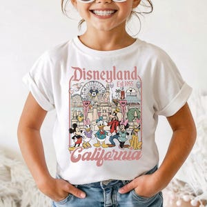 May include: A white short-sleeve t-shirt featuring a colorful graphic print of Disneyland California with Mickey Mouse, Minnie Mouse, Donald Duck, Daisy Duck, Goofy, and Pluto. The shirt is paired with a bright pink headband and round sunglasses.