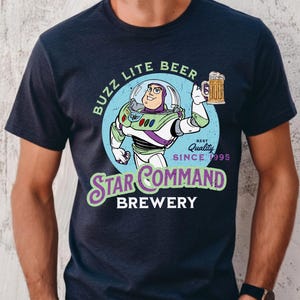 Puede incluir: Camiseta azul marino con un dise&ntilde;o de dibujos animados de Buzz Lightyear sosteniendo una jarra de cerveza. El dise&ntilde;o incluye el texto "Buzz Lite Beer" y "Star Command Brewery" con "Best Quality Since 1995". La camiseta es de cuello redondo.