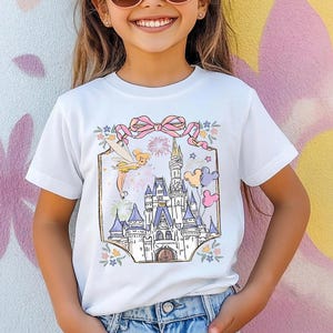 May include: White t-shirt featuring a colorful illustration of a fairytale castle with fireworks, a fairy, and balloons. The design is framed by a pink bow and floral accents. The shirt is worn by a young person with sunglasses.