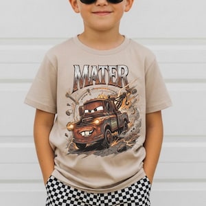 Puede incluir: Una camiseta beige de manga corta con un gr&aacute;fico del personaje Mater de la pel&iacute;cula Cars. La palabra "MATER" est&aacute; impresa encima de la imagen. El ni&ntilde;o lleva gafas de sol negras y pantalones cortos a cuadros.