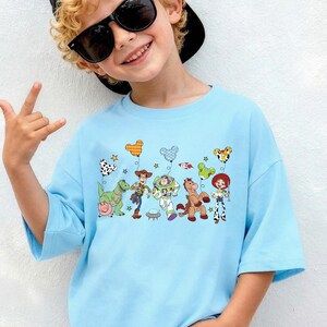 May include: Light blue t-shirt featuring a colorful graphic of Toy Story characters. The design includes Woody, Buzz Lightyear, and other characters from the movie. The shirt has short sleeves and a relaxed fit, perfect for casual wear.