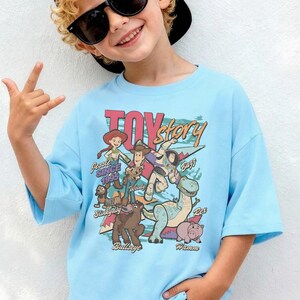 Puede incluir: Una camiseta azul claro con un estampado gr&aacute;fico colorido de los personajes de Toy Story, incluyendo a Woody, Buzz Lightyear, Jessie, Perdig&oacute;n, Slinky, Rex y Hamm, con el texto Toy Story y los nombres de los personajes en fuente retro.