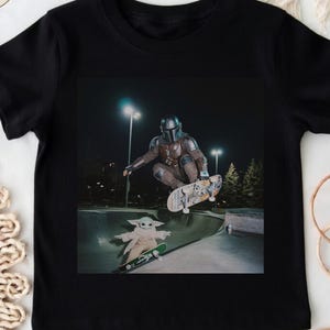 May include: Black t-shirt featuring a graphic of The Mandalorian and Grogu skateboarding in a skate park. The Mandalorian is mid-air on a skateboard, while Grogu is skateboarding below. The shirt has the text "SnuggleNestThreads" on the upper right.