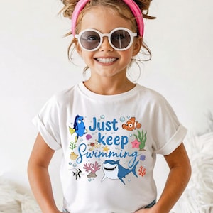 May include: A white short-sleeve t-shirt featuring a colorful graphic print of various sea creatures, including a blue tang, a clownfish, and a shark, with the text Just keep Swimming in blue script, paired with blue denim jeans.