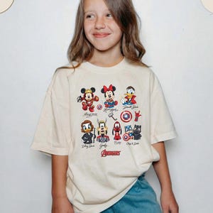 May include: A cream-colored t-shirt featuring cartoon characters, including Mickey Mouse, Minnie Mouse, and Donald Duck, with the word "Avengers" in red. The shirt is worn by a person.
