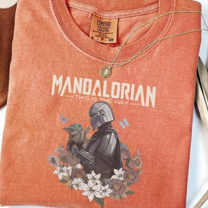Puede incluir: Una camiseta de manga corta color naranja quemado con un gr&aacute;fico del Mandaloriano y Grogu rodeados de flores y mariposas. El texto dice Mandalorian This Is The Way. La camiseta tiene cuello redondo y un acabado lavado estilo vintage.