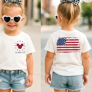 May include: A young child wearing a white t-shirt with a red, white, and blue design. The front of the shirt features a Mickey Mouse head and the name "Scarlet". The back of the shirt displays a distressed American flag. The child is also wearing denim shorts and heart-shaped sunglasses.