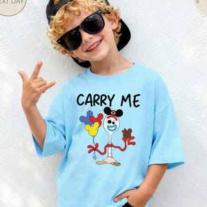 May include: A light blue t-shirt with the words "CARRY ME" in black. The shirt features a cartoon character holding balloons and a chocolate treat. The child is wearing sunglasses and a black cap.