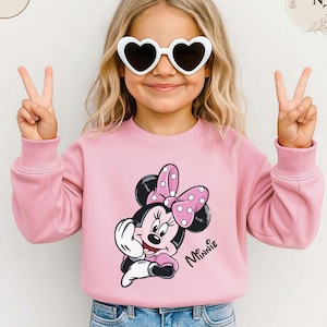 May include: A pink sweatshirt featuring a Minnie Mouse graphic with a polka dot bow. The word "Minnie" is written in cursive. The child is wearing white heart-shaped sunglasses and making a peace sign with their fingers.