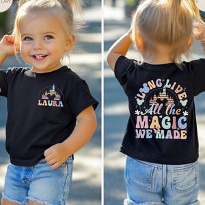 May include: Black t-shirt with a colorful castle graphic and the name "LAURA" on the front. The back of the shirt features the text "LONG LIVE All the MAGIC WE MADE" in pastel colors. The child is wearing denim shorts.
