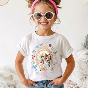 May include: A white t-shirt featuring a gold-framed portrait of Disney princesses surrounded by cartoon illustrations. The girl in the image is wearing sunglasses and a pink headband. The shirt is paired with blue jeans.