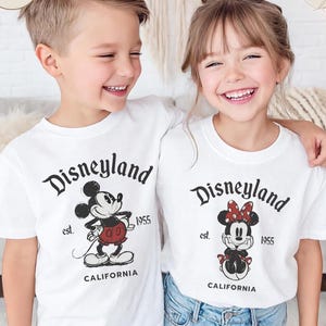 Pu&ograve; includere: Due bambini sorridenti indossano magliette bianche con una grafica di Disneyland. Le magliette presentano Topolino e Minnie, la scritta "Disneyland", "est. 1955" e "California".