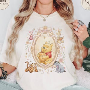 May include: Cream-colored t-shirt featuring a Winnie the Pooh design. The graphic includes Winnie the Pooh holding a honey pot, surrounded by other characters like Piglet, Tigger, Eeyore, and Rabbit. The shirt also has the text "SnuggleNestThreads" and "Ships Next Day".