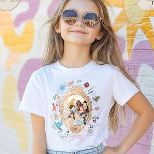 May include: A white t-shirt with a gold-framed illustration of Disney princesses. The design includes various characters, a red apple, and the word "Moana". The model is wearing round sunglasses.