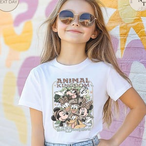 Pu&ograve; includere: T-shirt bianca con una grafica di personaggi Disney, tra cui Topolino e Minnie, Pippo e Paperino, con la scritta "ANIMAL KINGDOM" e "Walt Disney World". La maglietta &egrave; indossata da una persona giovane.