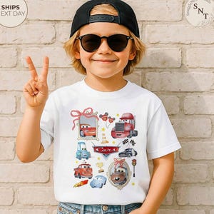 May include: White t-shirt featuring colorful watercolor illustrations of various Cars characters. The design includes images of Lightning McQueen, Mater, and other vehicles, along with the word "Cars" in a stylized font. The shirt is worn by a child.