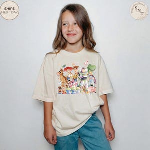 May include: A cream-colored t-shirt featuring a colorful graphic of characters from the Toy Story movie. The design includes Woody, Buzz Lightyear, Jessie, and other beloved characters. The shirt is worn by a young person.