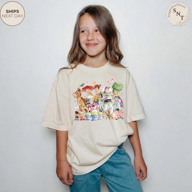 May include: A cream-colored t-shirt featuring a colorful graphic of characters from the Toy Story movie. The design includes Woody, Buzz Lightyear, Jessie, and other beloved characters. The shirt is worn by a young person.