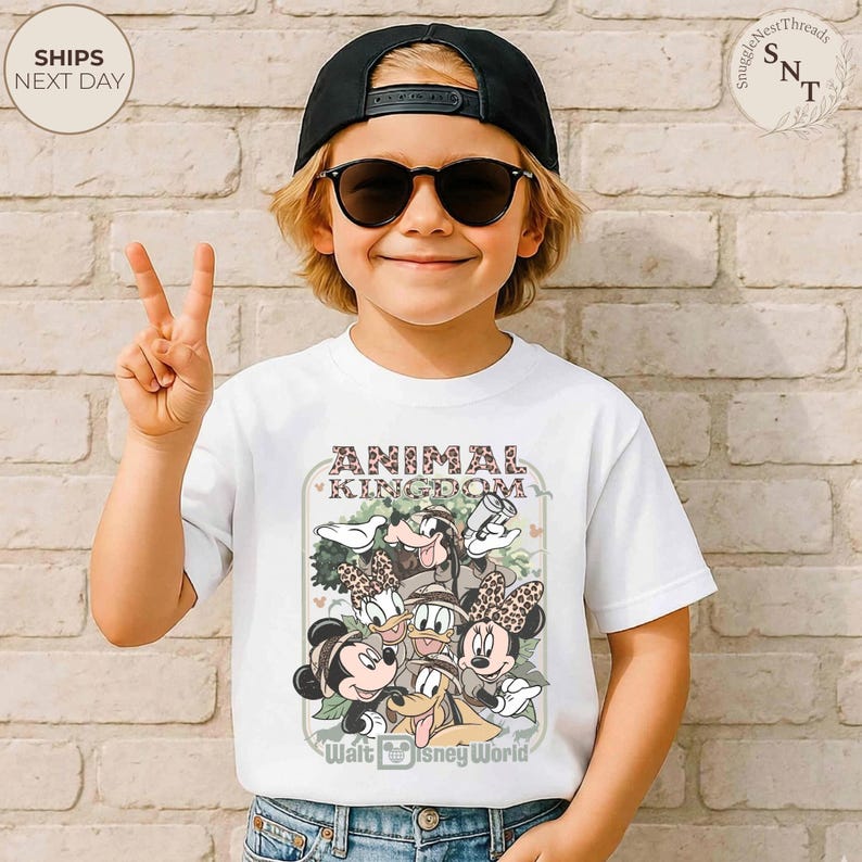 Pu&ograve; includere: T-shirt bianca con una grafica dei personaggi Disney, tra cui Topolino e Minnie, Paperino e Pippo, con la scritta "Animal Kingdom" e "Walt Disney World". La maglietta &egrave; indossata da un bambino con un berretto nero e occhiali da sole.