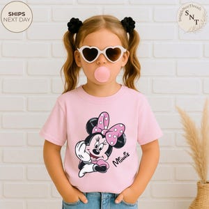 May include: A pink t-shirt featuring a Minnie Mouse graphic with a polka dot bow. The shirt is worn by a child wearing heart-shaped sunglasses and has black hair ties. The words "Minnie" are written on the shirt.