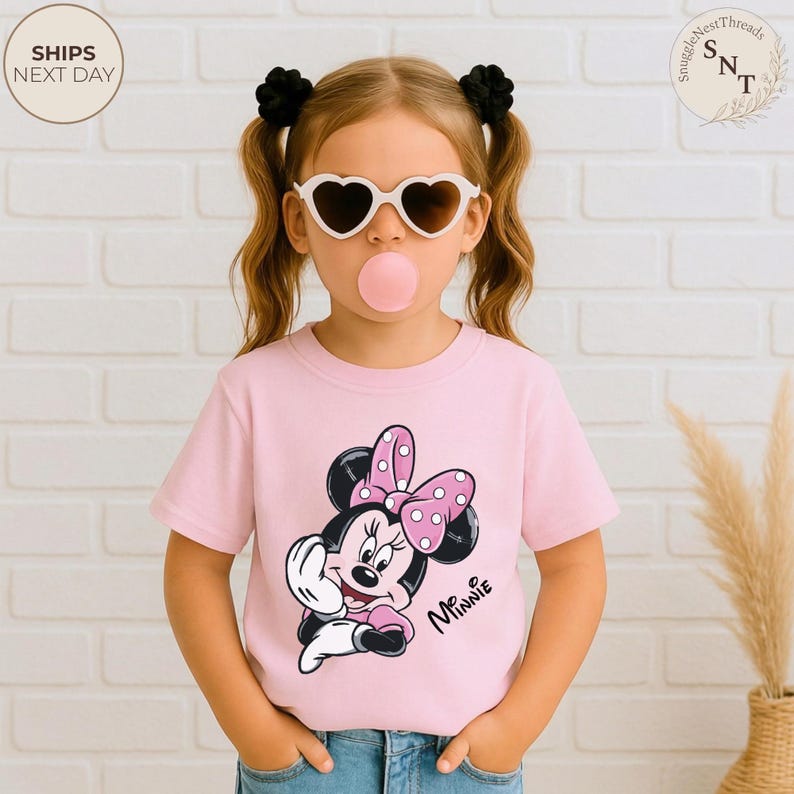 May include: A pink t-shirt featuring a Minnie Mouse graphic with a polka dot bow. The shirt is worn by a child wearing heart-shaped sunglasses and has black hair ties. The words "Minnie" are written on the shirt.