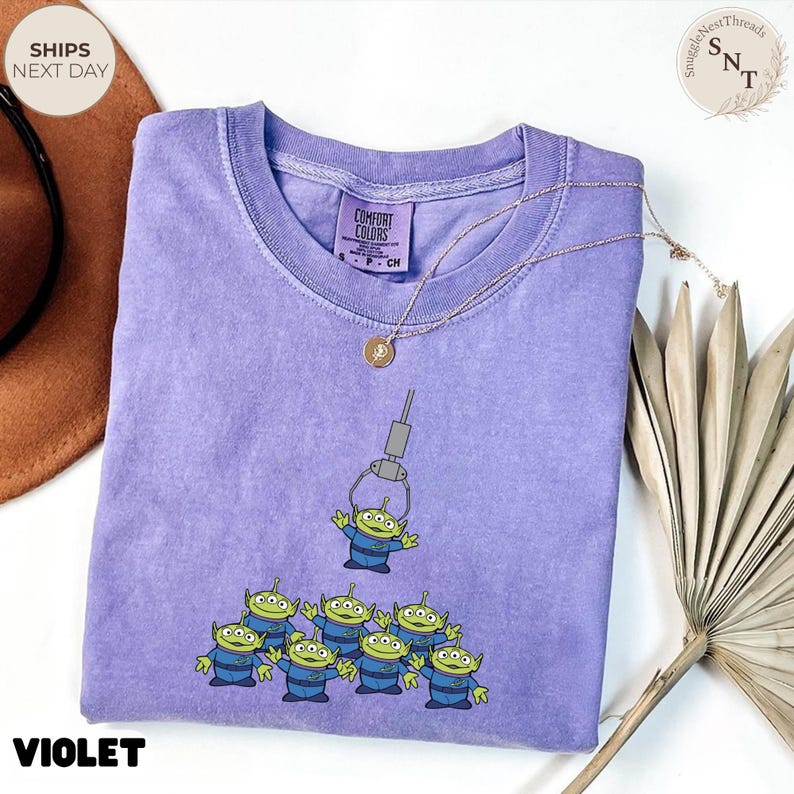 May include: Violet Comfort Colors t-shirt featuring a graphic of green aliens and a claw machine. The design includes the word "VIOLET" at the bottom. A gold necklace with a pendant is draped over the shirt.