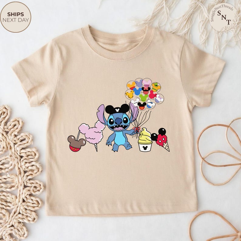 Pu&ograve; includere: T-shirt beige con un personaggio dei cartoni animati blu con un cappello nero di Topolino, che tiene in mano palloncini e dolcetti. La maglietta ha il testo "SHIPS NEXT DAY" in alto a sinistra.