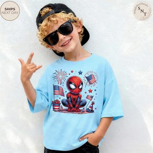 May include: A light blue t-shirt featuring a graphic of a cartoon Spider-Man character surrounded by American flags, fireworks, and patriotic party decorations. The design includes red, white, and blue elements, with the character centered on the chest.