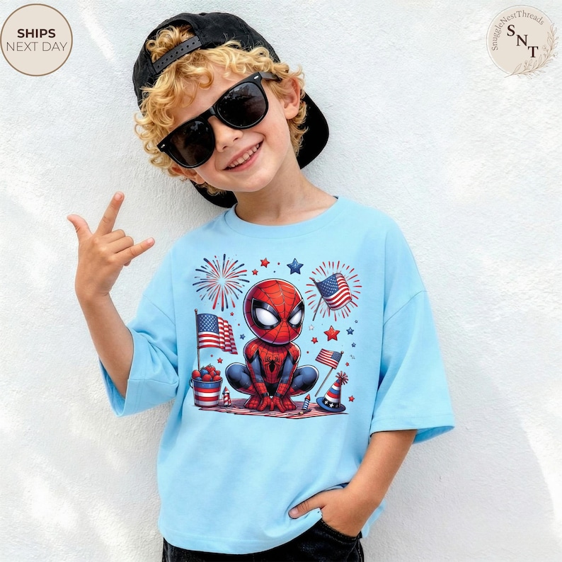 May include: A light blue t-shirt featuring a graphic of a cartoon Spider-Man character surrounded by American flags, fireworks, and patriotic party decorations. The design includes red, white, and blue elements, with the character centered on the chest.