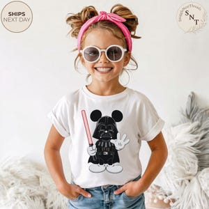 May include: White t-shirt featuring a cartoon Darth Vader character with Mickey Mouse ears, holding a red lightsaber and waving. The shirt has rolled-up sleeves. The image includes the text "SHIPS NEXT DAY" in a circle.