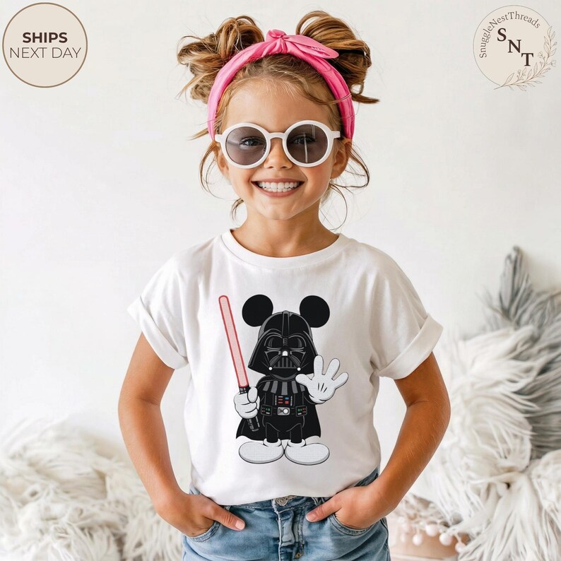 May include: White t-shirt featuring a cartoon Darth Vader character with Mickey Mouse ears, holding a red lightsaber and waving. The shirt has rolled-up sleeves. The image includes the text "SHIPS NEXT DAY" in a circle.