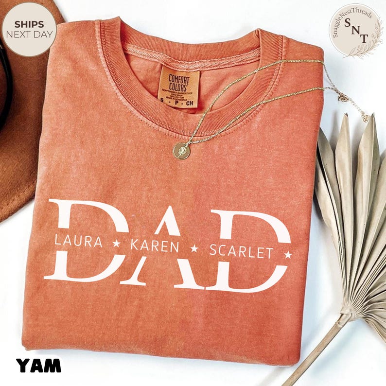 Puede incluir: Una camiseta de color coral con la palabra "DAD" impresa en letras blancas grandes. Debajo de la palabra "DAD" est&aacute;n los nombres "LAURA", "KAREN" y "SCARLET" con estrellas. Un collar dorado con un colgante est&aacute; sobre la camiseta.