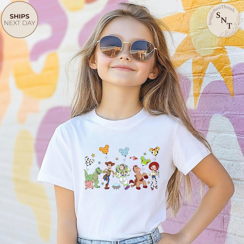 May include: White t-shirt featuring a colorful graphic of animated characters, including a dinosaur, cowboy, and space ranger. The design includes balloons and stars. The shirt is worn by a person wearing sunglasses.