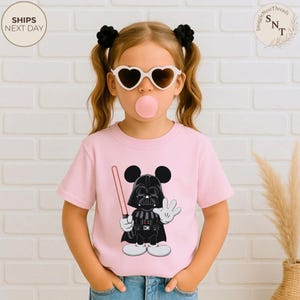 May include: Pink t-shirt featuring a cartoon character mashup of Darth Vader and Mickey Mouse, holding a red lightsaber and waving. The design is in black, white, and red. The shirt is worn by a child with heart-shaped sunglasses and a bubblegum bubble.