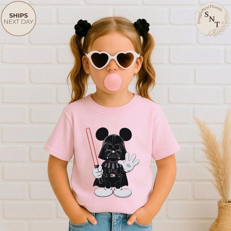 May include: Pink t-shirt featuring a cartoon character mashup of Darth Vader and Mickey Mouse, holding a red lightsaber and waving. The design is in black, white, and red. The shirt is worn by a child with heart-shaped sunglasses and a bubblegum bubble.