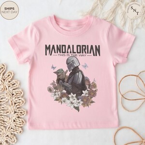 Puede incluir: Una camiseta de manga corta de color rosa claro con un gr&aacute;fico de The Mandalorian y Grogu rodeados de flores y mariposas. El texto "MANDALORIAN" y "THIS IS THE WAY" est&aacute; impreso en negro sobre los personajes en una fuente negrita.