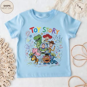 May include: Light blue t-shirt featuring the words "TOY STORY" in colorful letters. The shirt showcases a vibrant graphic of characters from the animated film, including Woody, Buzz Lightyear, and Rex, with scribbled background details.